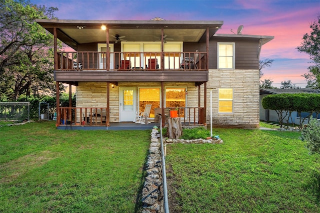 back house at dusk with a balcony, ceiling fan, a patio area, and a yard