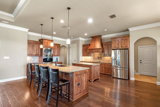 kitchen featuring premium range hood, a center island with sink, hanging light fixtures, dark hardwood / wood-style floors, and appliances with stainless steel finishes