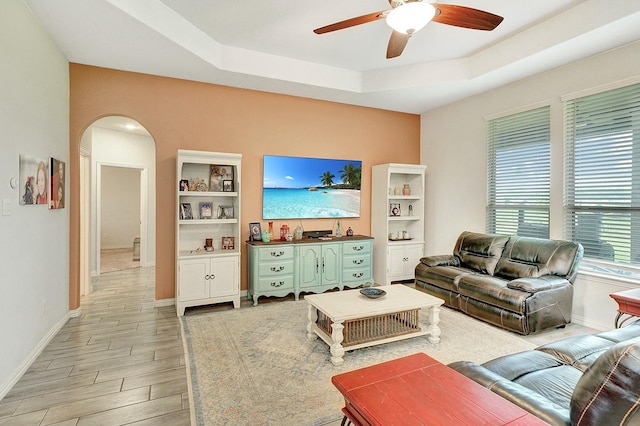 living area featuring arched walkways, ceiling fan, baseboards, light wood-type flooring, and a raised ceiling