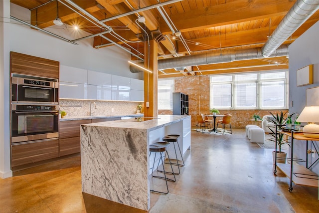 kitchen with light stone counters, brick wall, decorative backsplash, white cabinetry, and double oven