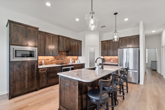 kitchen with a kitchen island with sink, light stone countertops, light wood-type flooring, dark brown cabinetry, and stainless steel appliances