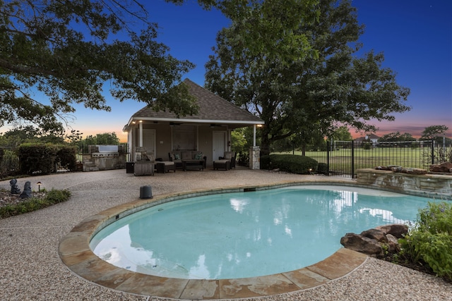 pool at dusk featuring exterior kitchen, ceiling fan, an outdoor living space, and a patio