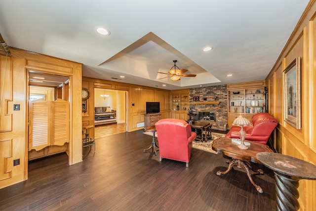 living room with a brick fireplace, ceiling fan, a raised ceiling, dark wood-type flooring, and built in shelves