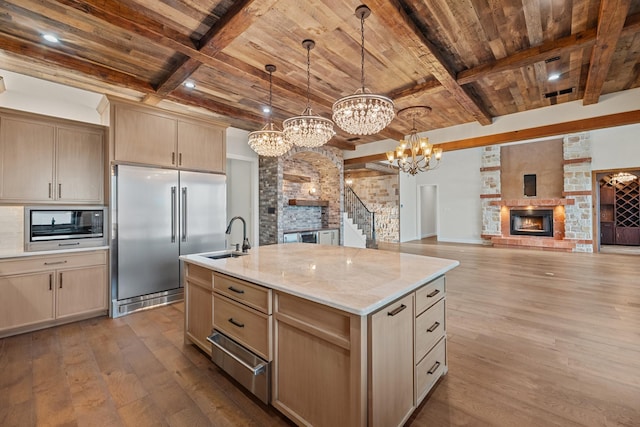 kitchen featuring light brown cabinetry, decorative light fixtures, a fireplace, an island with sink, and built in appliances