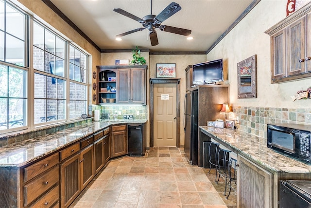 kitchen with light stone counters, black appliances, a kitchen breakfast bar, ceiling fan, and ornamental molding