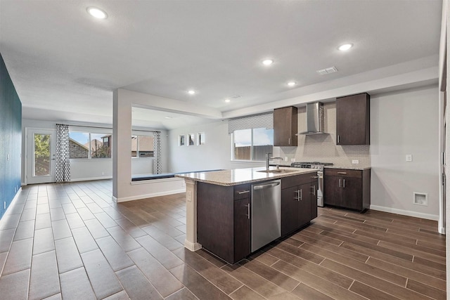 kitchen featuring wall chimney range hood, sink, an island with sink, appliances with stainless steel finishes, and dark brown cabinetry