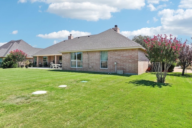 back of property with roof with shingles, brick siding, a chimney, a patio, and a lawn