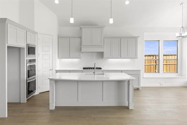 kitchen with decorative light fixtures, a center island with sink, high vaulted ceiling, light wood-type flooring, and stainless steel microwave