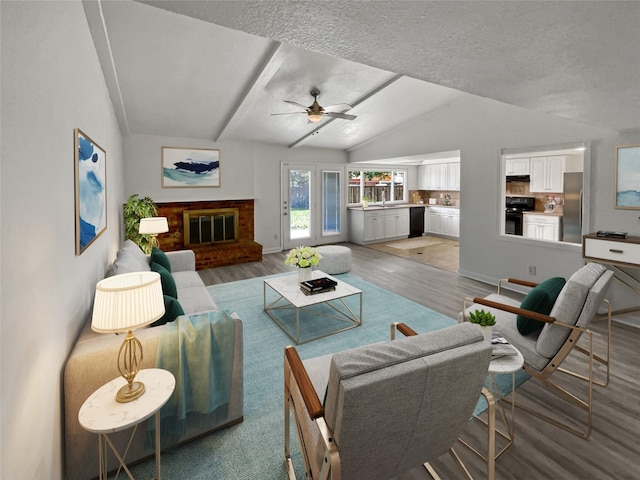 living room with light wood-type flooring, vaulted ceiling with beams, ceiling fan, a brick fireplace, and a textured ceiling