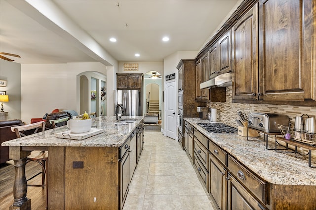 kitchen with a kitchen island with sink, decorative backsplash, ceiling fan, light stone counters, and stainless steel appliances