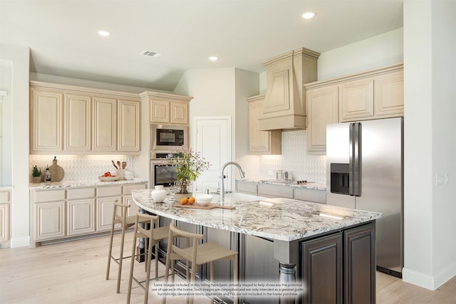 kitchen with a kitchen island with sink, sink, custom exhaust hood, appliances with stainless steel finishes, and light hardwood / wood-style floors