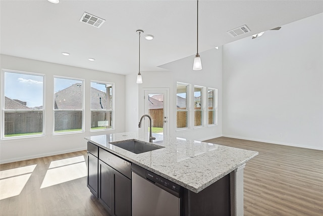 kitchen with stainless steel dishwasher, hanging light fixtures, sink, light stone counters, and an island with sink