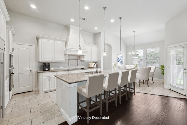 kitchen featuring sink, premium range hood, white cabinetry, light stone counters, and a center island with sink