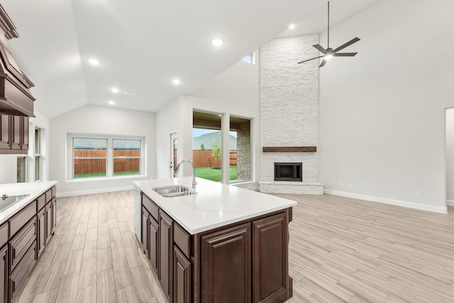 kitchen featuring a stone fireplace, a kitchen island with sink, sink, ceiling fan, and light wood-type flooring