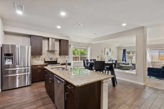 kitchen with a kitchen island with sink, dark brown cabinets, stainless steel appliances, and wall chimney range hood