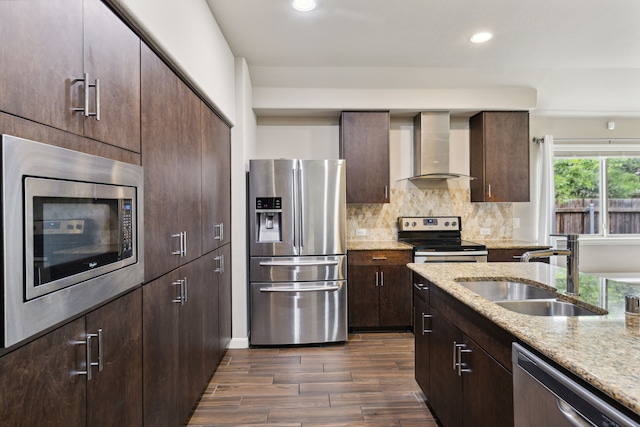 kitchen with light stone countertops, appliances with stainless steel finishes, dark wood-type flooring, sink, and wall chimney range hood