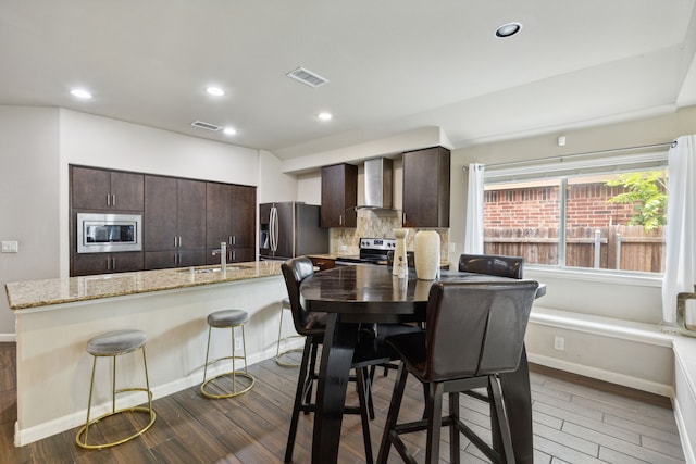 kitchen featuring a breakfast bar area, dark brown cabinets, dark hardwood / wood-style flooring, stainless steel appliances, and wall chimney range hood