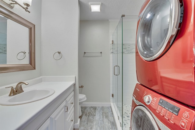 bathroom with stacked washing maching and dryer, a textured ceiling, toilet, vanity, and a shower with shower door