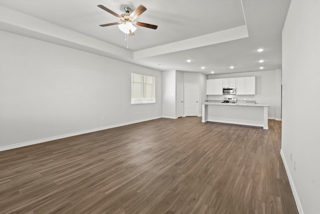 unfurnished living room with baseboards, dark wood finished floors, a ceiling fan, a tray ceiling, and recessed lighting