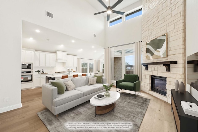 living room with ceiling fan, light hardwood / wood-style floors, a stone fireplace, and a high ceiling