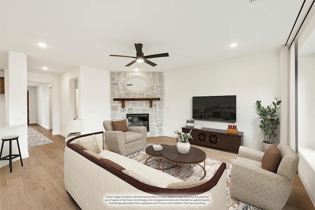 living room with light wood-type flooring, a stone fireplace, and ceiling fan