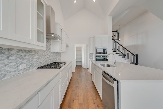 kitchen with appliances with stainless steel finishes, high vaulted ceiling, wall chimney range hood, white cabinets, and sink