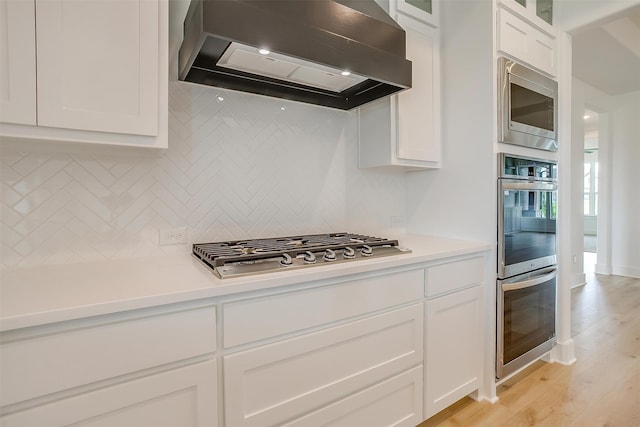 kitchen with appliances with stainless steel finishes, tasteful backsplash, custom exhaust hood, light wood-type flooring, and white cabinets