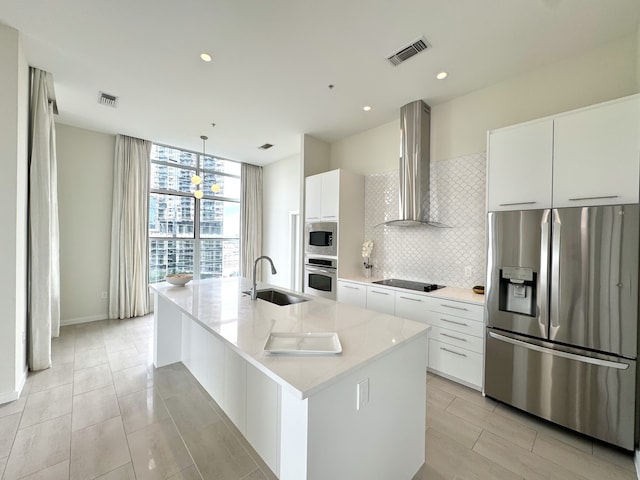 kitchen featuring stainless steel appliances, backsplash, wall chimney range hood, sink, and a kitchen island with sink