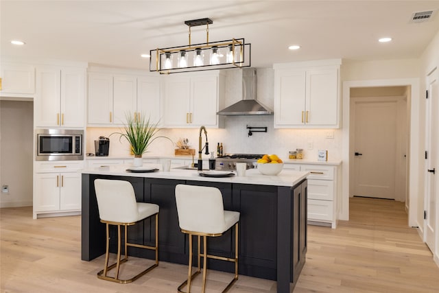 kitchen with wall chimney exhaust hood, stainless steel microwave, white cabinetry, and a kitchen island with sink