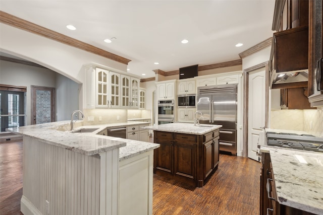 kitchen featuring sink, light stone counters, built in appliances, kitchen peninsula, and an island with sink