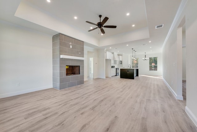 unfurnished living room featuring a tiled fireplace, ceiling fan with notable chandelier, a raised ceiling, and light hardwood / wood-style flooring