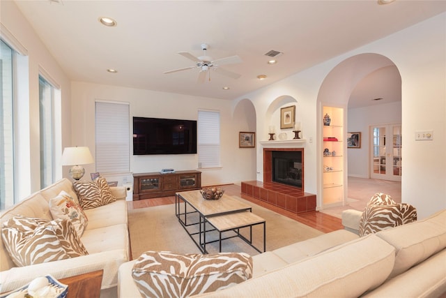 living room featuring built in shelves, ceiling fan, light wood-type flooring, and a tiled fireplace