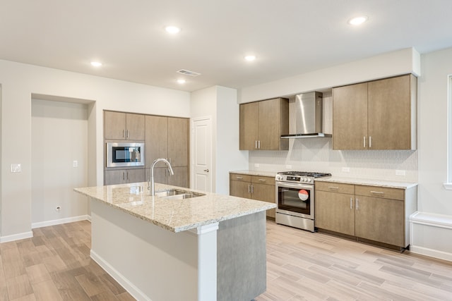 kitchen with light hardwood / wood-style floors, a center island with sink, sink, appliances with stainless steel finishes, and wall chimney exhaust hood