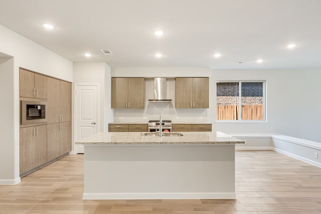 kitchen featuring sink, an island with sink, wall chimney range hood, light stone countertops, and light hardwood / wood-style flooring