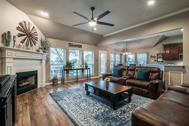 living room featuring vaulted ceiling, ceiling fan with notable chandelier, a tiled fireplace, crown molding, and dark wood-type flooring