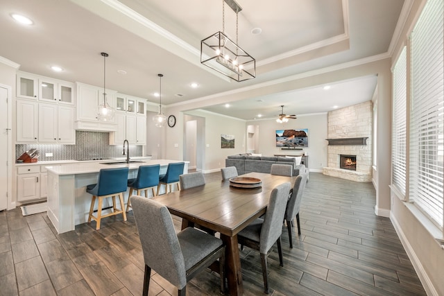 dining space featuring a stone fireplace, sink, crown molding, ceiling fan, and a tray ceiling