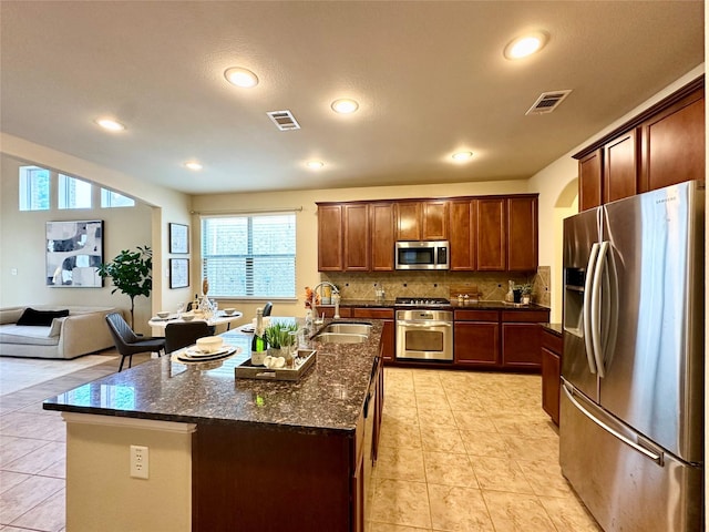 kitchen with sink, decorative backsplash, dark stone countertops, an island with sink, and stainless steel appliances