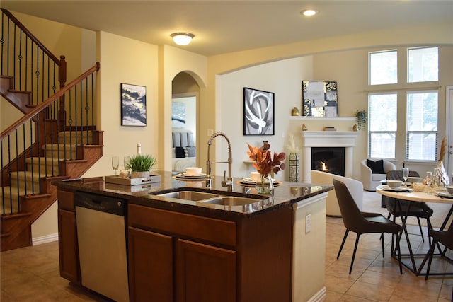 kitchen featuring stainless steel dishwasher, sink, light tile patterned floors, dark stone countertops, and an island with sink