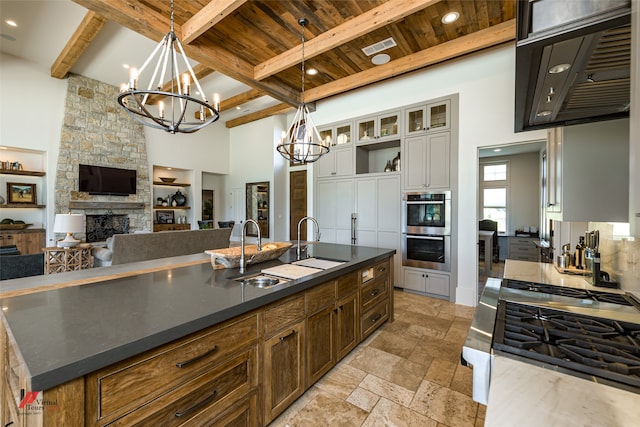 kitchen with beamed ceiling, a stone fireplace, sink, an island with sink, and double oven