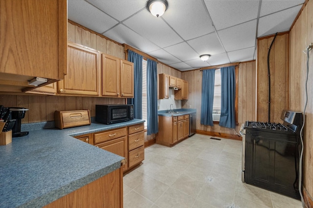 kitchen featuring a drop ceiling, black range with gas stovetop, wooden walls, sink, and dishwasher