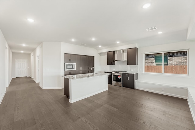 kitchen with hardwood / wood-style flooring, wall chimney exhaust hood, stainless steel appliances, a center island with sink, and light stone countertops