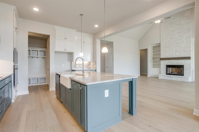 kitchen featuring a stone fireplace, an island with sink, sink, pendant lighting, and white cabinets