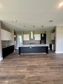 kitchen with white cabinetry, wall chimney exhaust hood, hardwood / wood-style flooring, and a kitchen island with sink