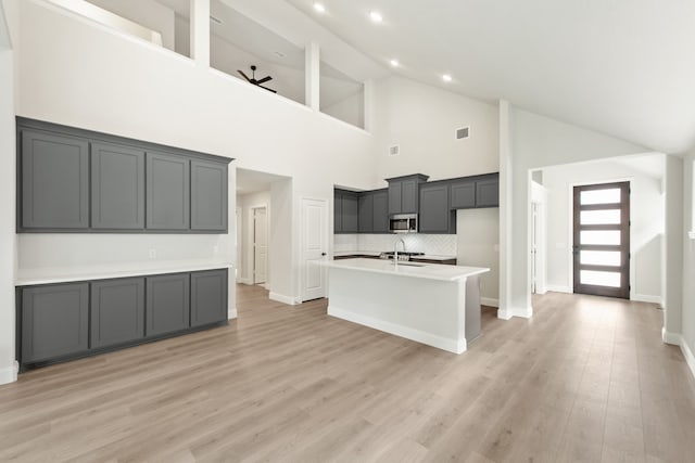 kitchen with gray cabinetry, light hardwood / wood-style floors, an island with sink, and high vaulted ceiling