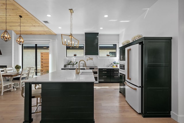 kitchen with light stone counters, hanging light fixtures, a kitchen island with sink, and high end fridge