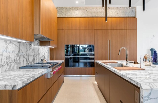 kitchen with sink, light stone countertops, an island with sink, double oven, and stainless steel gas cooktop