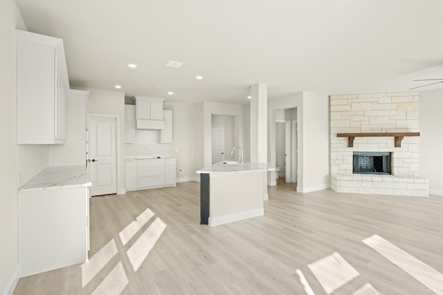 kitchen featuring white cabinets, decorative backsplash, a center island with sink, light hardwood / wood-style flooring, and a stone fireplace