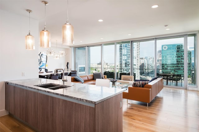 kitchen with light wood-type flooring, light stone counters, sink, a wall of windows, and hanging light fixtures