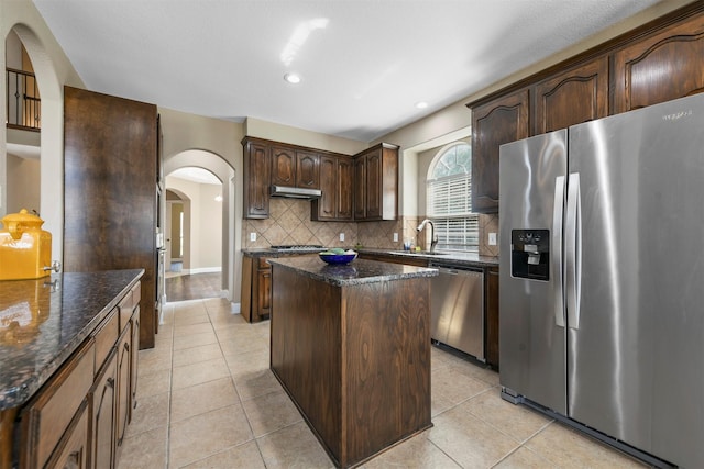 kitchen featuring dark brown cabinetry, a center island, tasteful backsplash, dark stone counters, and appliances with stainless steel finishes