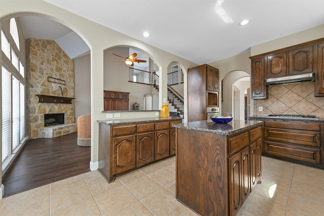 kitchen featuring a center island, tasteful backsplash, range hood, dark stone countertops, and a fireplace